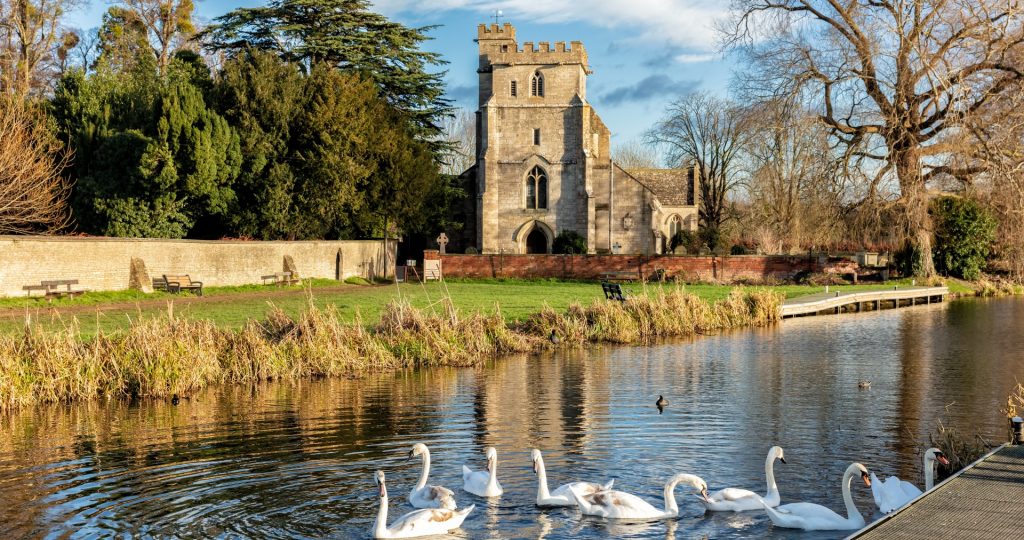Group of swans on lake in forefront, with medieval church in the background. church is Saint Cyr's Church in Gloucestershire. What's on in Gloucestershire, Cheltenham, Stroud. 